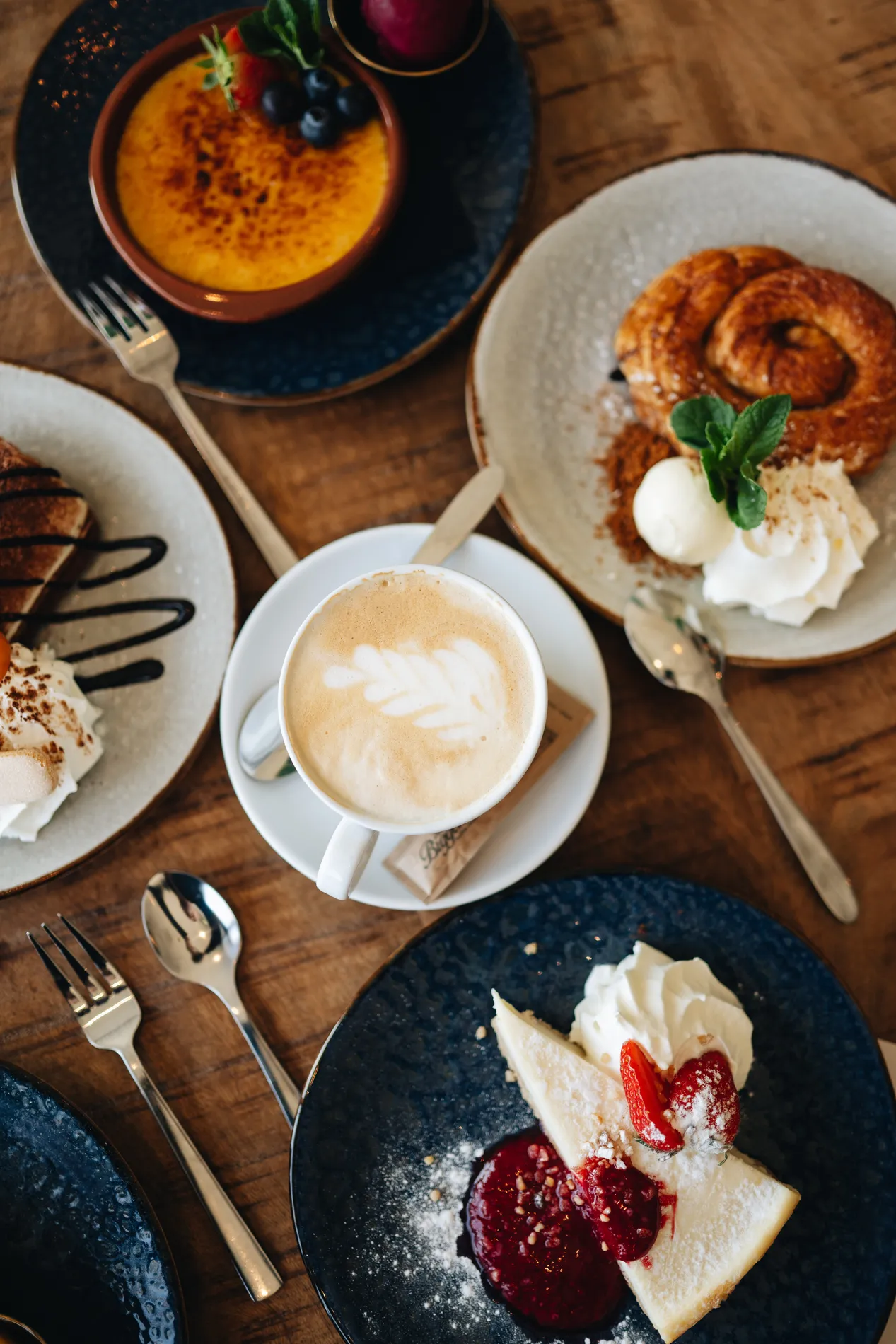 Bovenaanzicht van een koffie- en desserttafel met in het midden een cappuccino met latte art, omringd door cheesecake met rood fruit, crème brûlée en een zoet bladerdeeggebakje met ijs.